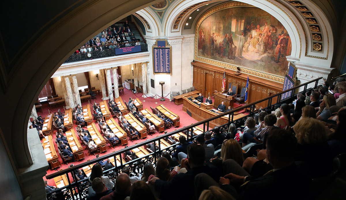 a photo of the Assembly Chamber from the balcony showing people seated in the balcony and in seats on the floor and at the head of the room