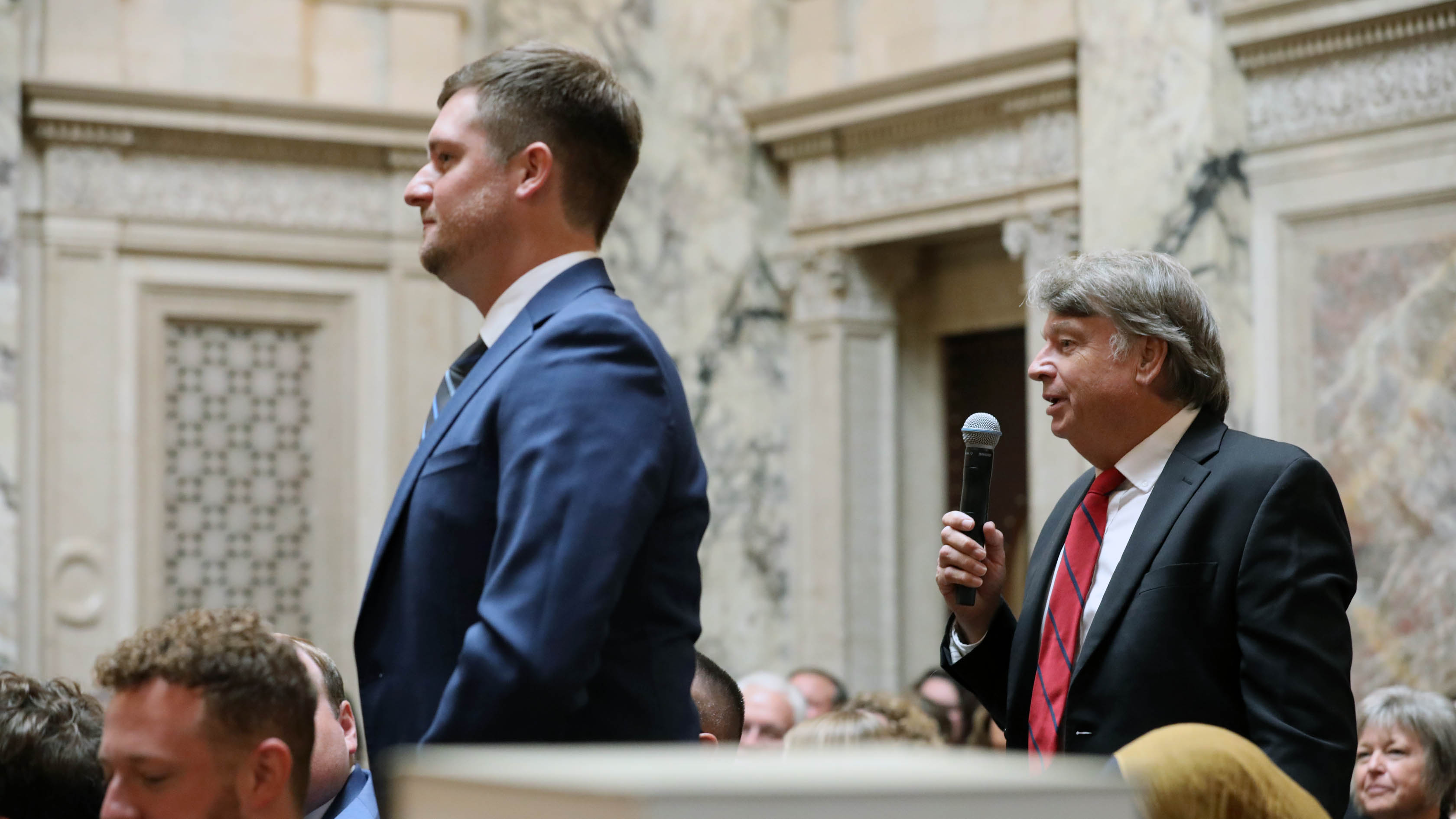 two men standing among a seated group with one holding a microphone