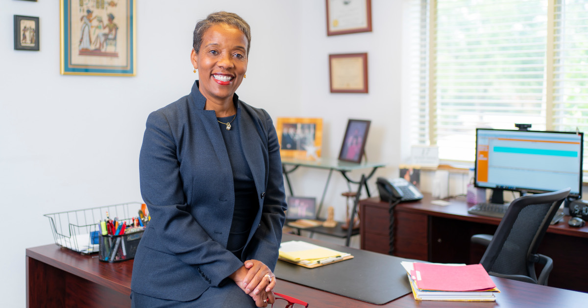 Michelle Behnke, A Middle Aged Black Woman A BLue-Gray Business Suit, Suits On The Side Of The Desk In Her Solo Law Office