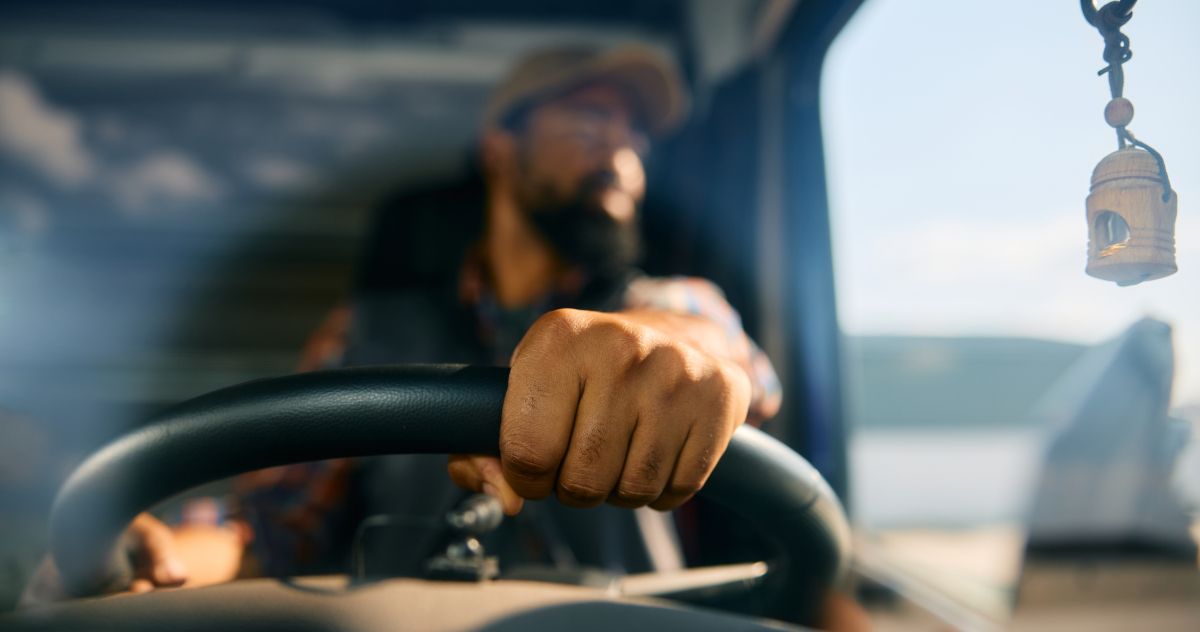 Close Up Of A Black Man's Hands On The Wheel Of A Tractor Trailer, With The Man's Upper Body, His Head Turned Toward The Cab's Window, Blurred By Shallow Depth Of Field