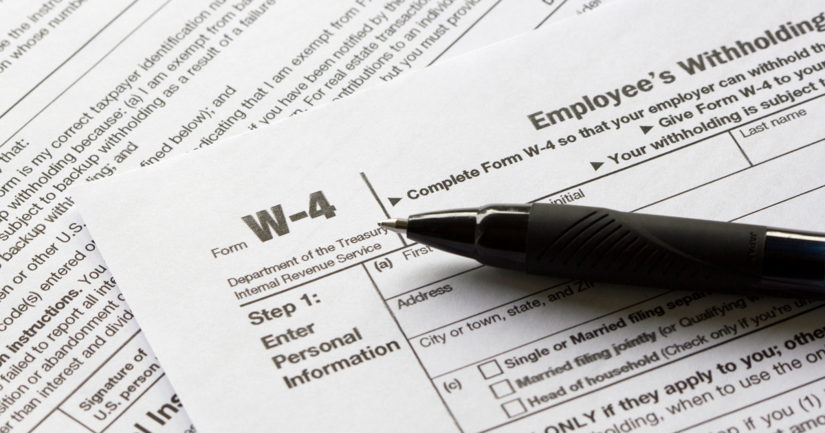 A W-4 Employee Withholding Form Lying Atop Another Form, With A Black Ball Point Pen Lying At An Angle Across The W-4 Form