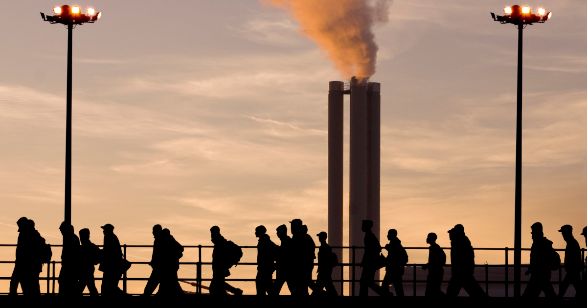 Silhouetted Against A Blue Sky Painted Orange By The Setting Sun, A Line Of Factory Workers Leave Work Along A Gangplank, With Two Belching Smokestacks In The Background