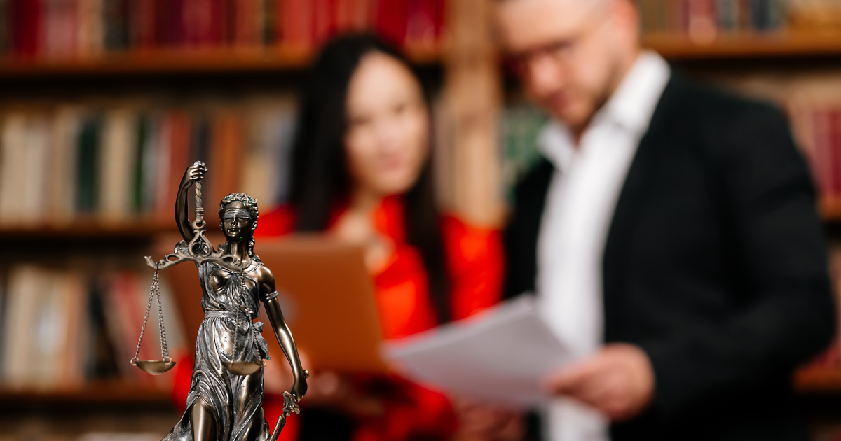 Close Up Of A Small Silver Statue of Lady Justice; In The Medium Distance, Artfully Out Of Foces, A Woman In A Red Dress Holds A File Folder While Conferring With A Man In A Suit Standing Next to Her, With Bookshelves Filling The Background