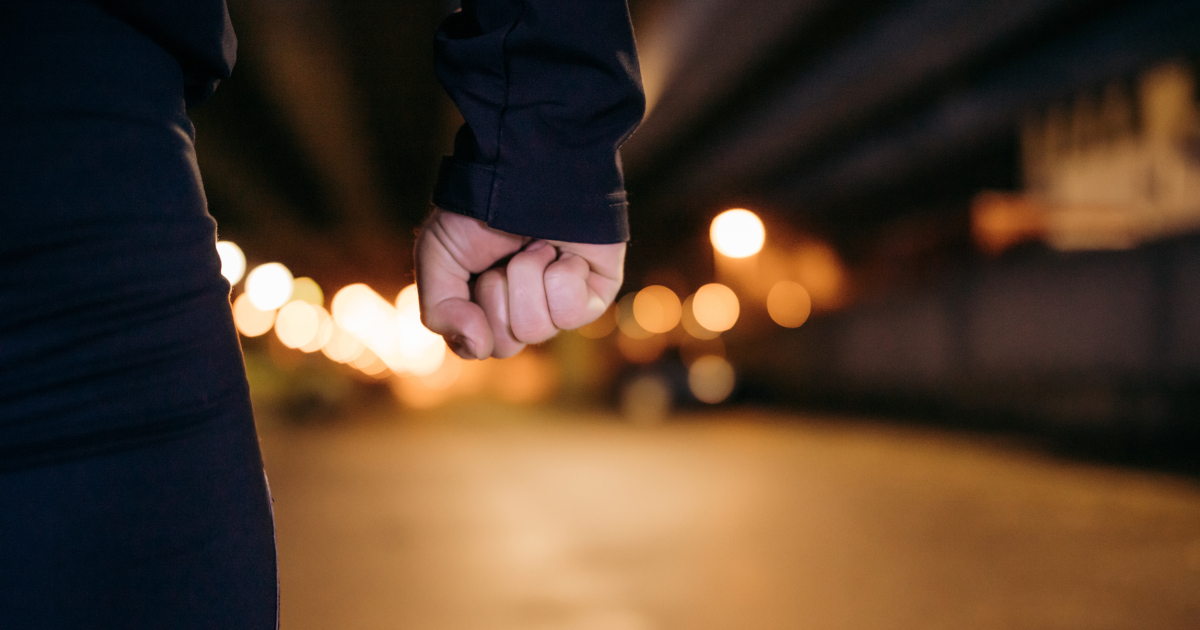 Close Up Of A Man's Balled Up Fist, His Arm In A Black Hoodie Hanging Down Next To His Jeans-Clad Thigh, With Blurry Streetlights Glowing Cinematically At Night In The Distance