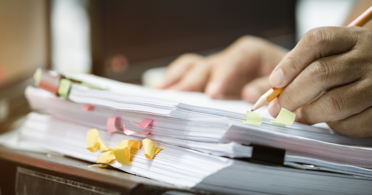 Close Up Of A Pile Of Legal Papers, Marked With Crumpled Post It Notes, With a Person's Hand Holding A Pencil Above The Top Paper