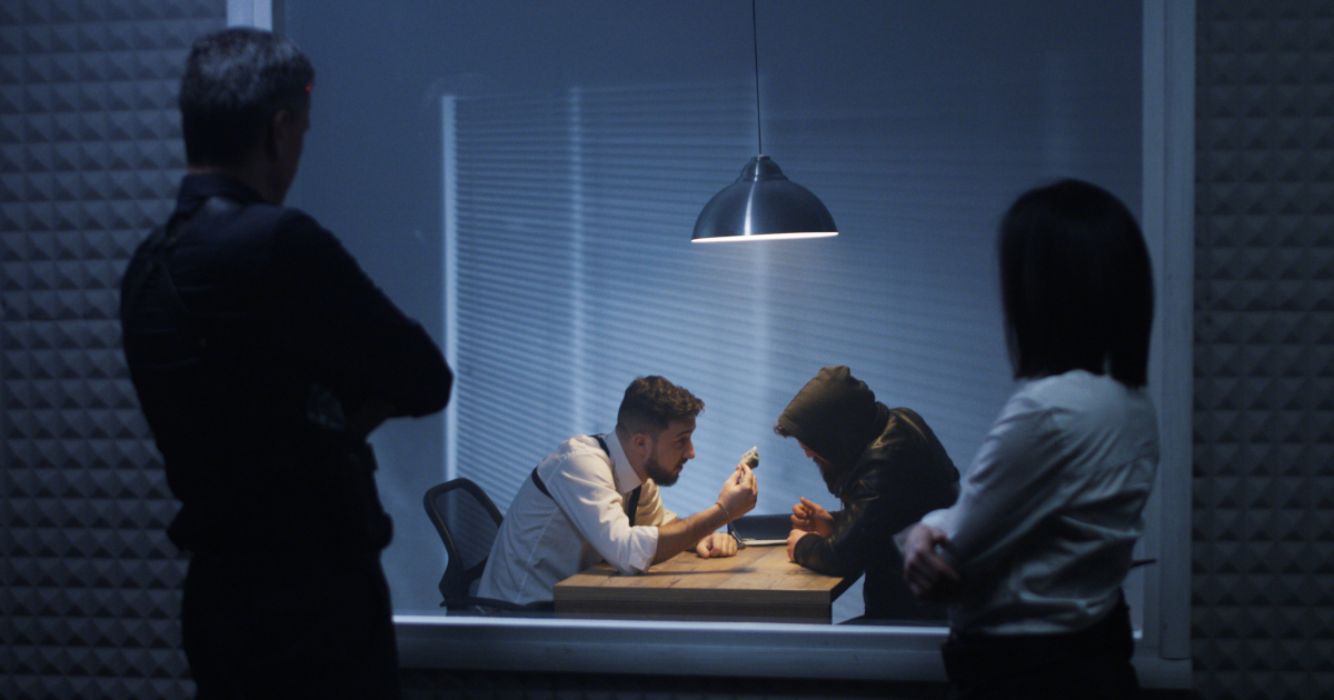 A Man And A Woman, Shrouded In Shadow And Standing Arms Akimbo, Standing In Front Of A One-Way Glass Panel, Watching Through The Glass As A Male Detective Intensely Questioning A Suspect Inside An Interrogation Room 