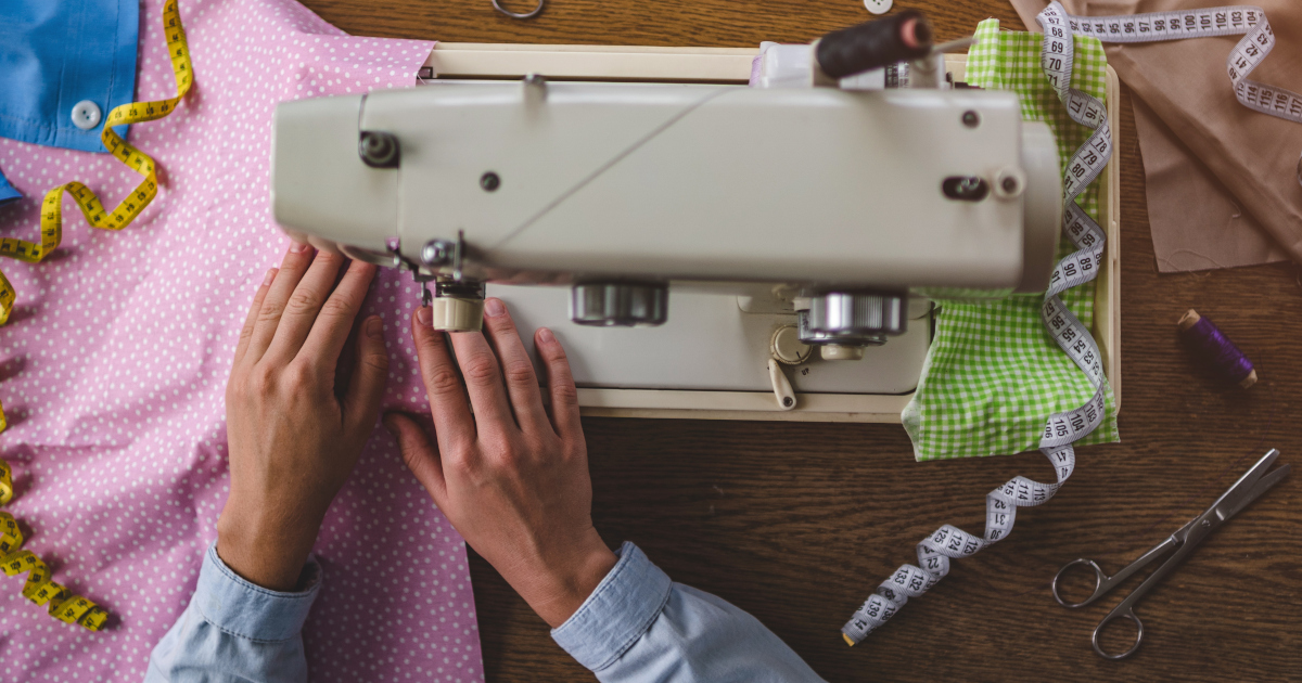Seen From Directly Overhead, A Pair of Hands Guides A Polka-Dottd Piece of Pink Fabric Beneath The Presser Foot Of A Sewing Machine, With Measuring Tapes, Scissors, And Spools Of Thread Lying On The Table Around The Sewing Machine
