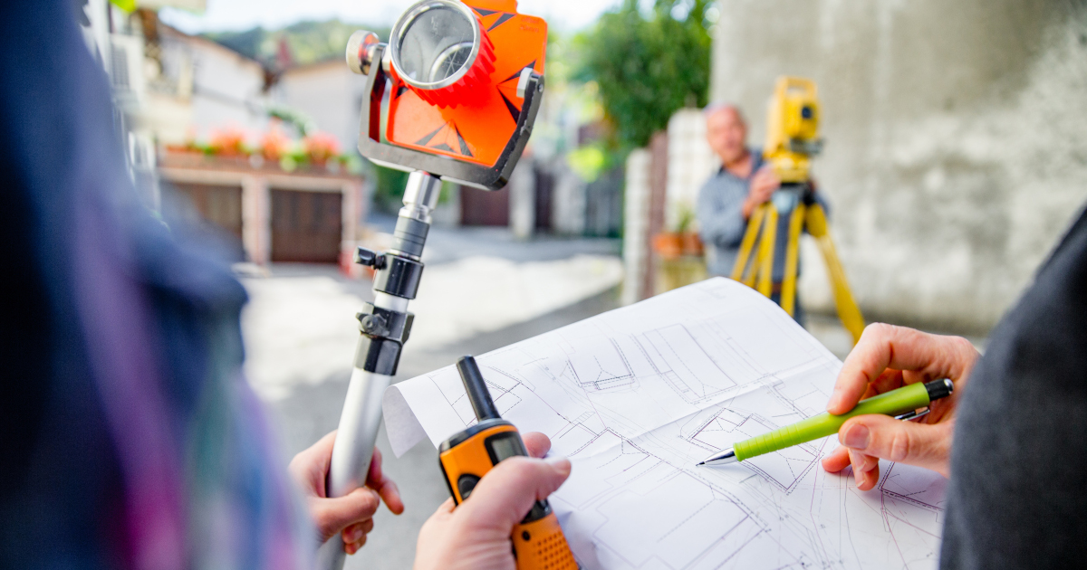 Closeup Of A Worker, On The Left, Holding A Surveying Tool On A Monopod, While On The Right A Man In A Sportcoat And Holding a Walkie Talkie In His Left Hand Points To A Survey Map With A Pen In His Right Hand, In The Distance A Surveyor Stands Behind A Yellow Survey Tool Mounted On A Tripod