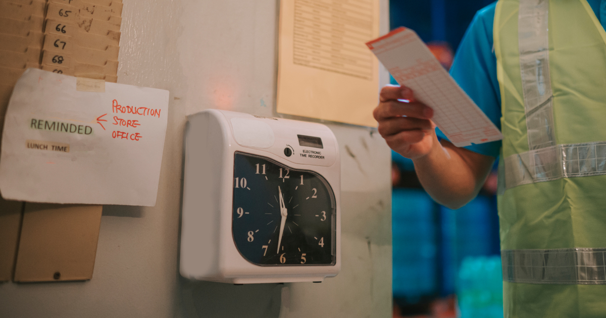 Close Up Of A Facory Punch Clock, With A Male Worker In A High Visibility Vest Standing To The Right Of The Clock With His Timecard In His Hand